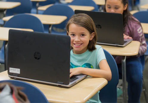 A young student smiling from behind her laptop.