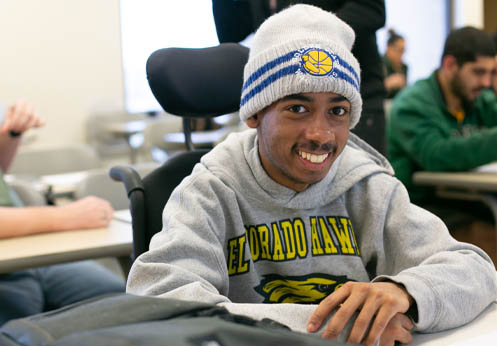 A smiling black male student sitting in his wheel chair at his desk