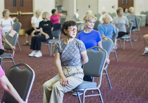 workout class with older students sitting on chairs performing stretches.