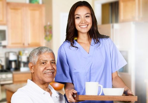 Young female personal care aide in blue scrubs serving coffee to elderly man at home, demonstrating caregiving skills.